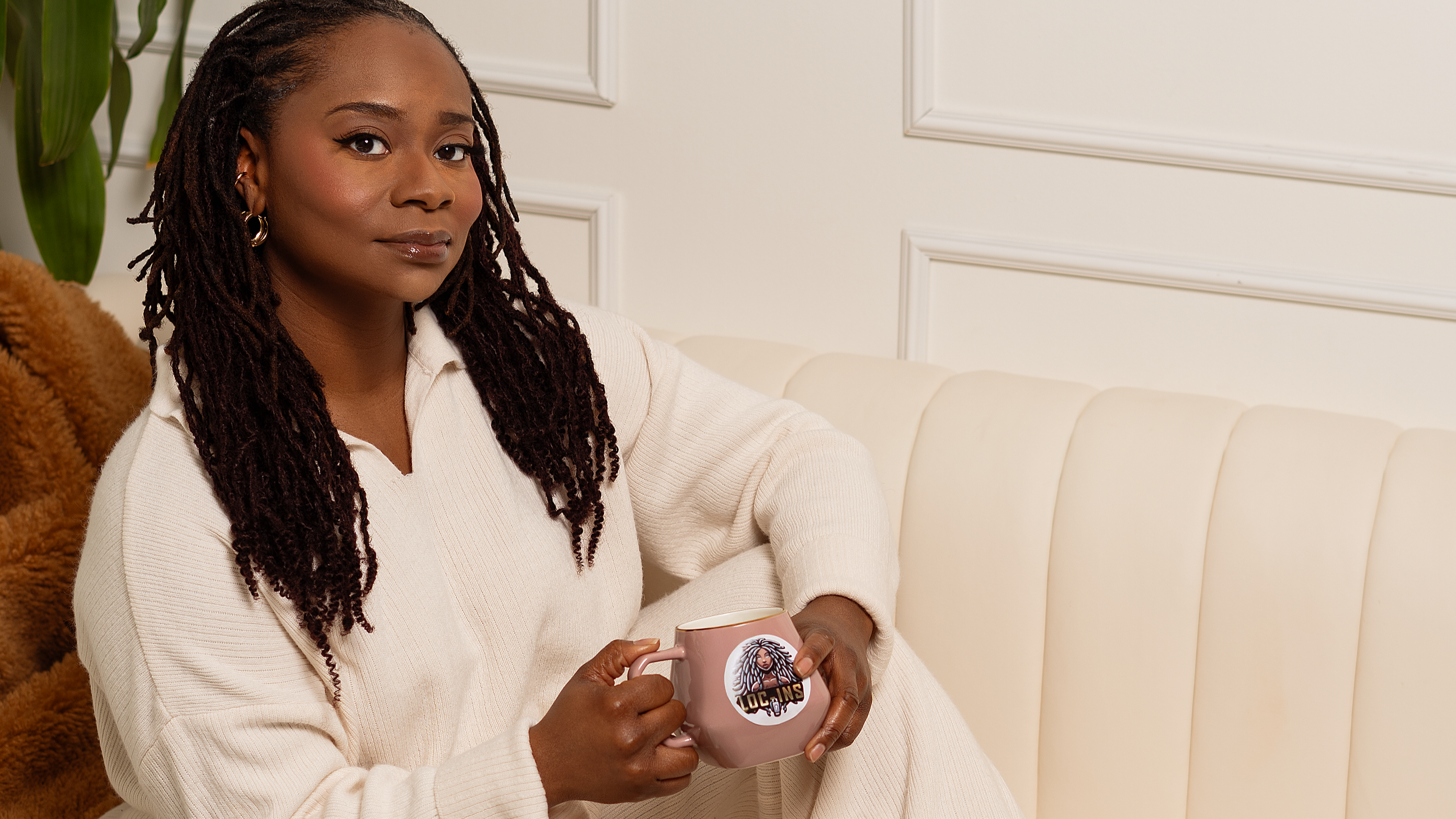 Loc-Ins model with dreadlocks in her hair sitting on a couch holding a pink coffee cup with the Loc-Ins logo on the mug.